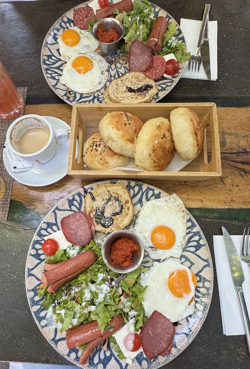 Two Bon Bouquet breakfast plates with fried eggs, sausages, salad and a pancake, with a basket of warm bread rolls and a cappuccino